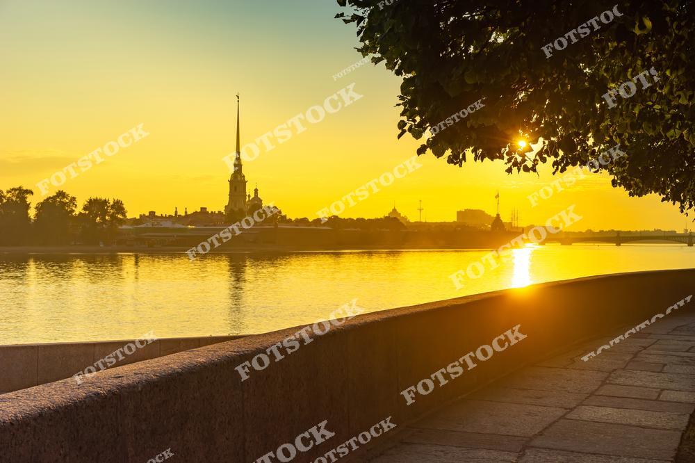 На фотографии изображена Петропавловская крепость на фоне закатного неба, отражающегося в воде Невы. Вид с набережной создает спокойную и романтичную атмосферу. Солнечные лучи проникают сквозь листву деревьев, добавляя теплых оттенков городскому пейзажу. Эта сцена отражает величие и красоту исторического Санкт-Петербурга.